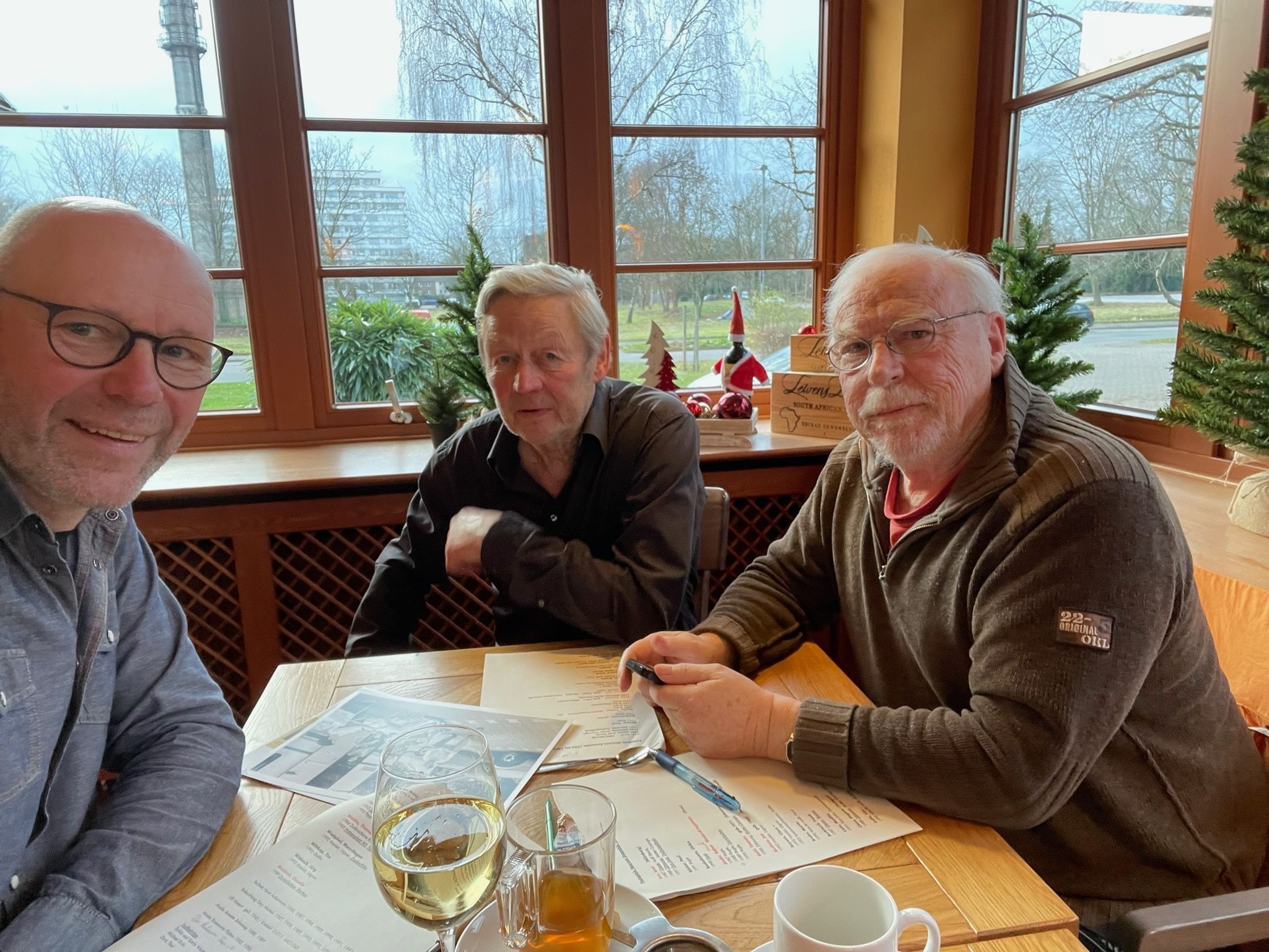 Drei Männer sitzen lächelnd in gemütlichem Restaurant mit Getränken und Blick ins Freie.
