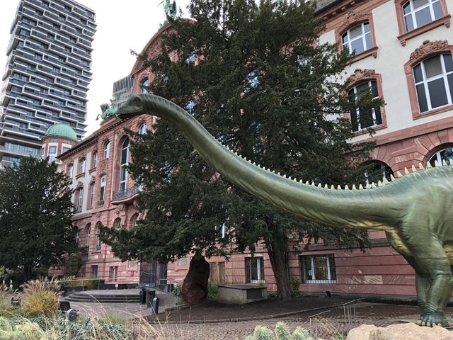 Lebensgroße Dinosaurier-Skulptur vor historischem Gebäude mit modernem Hochhaus im Hintergrund.