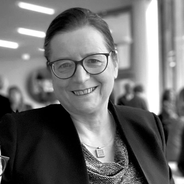 Lächelnde Person mit Brille in eleganter Jacke bei einer Veranstaltung.